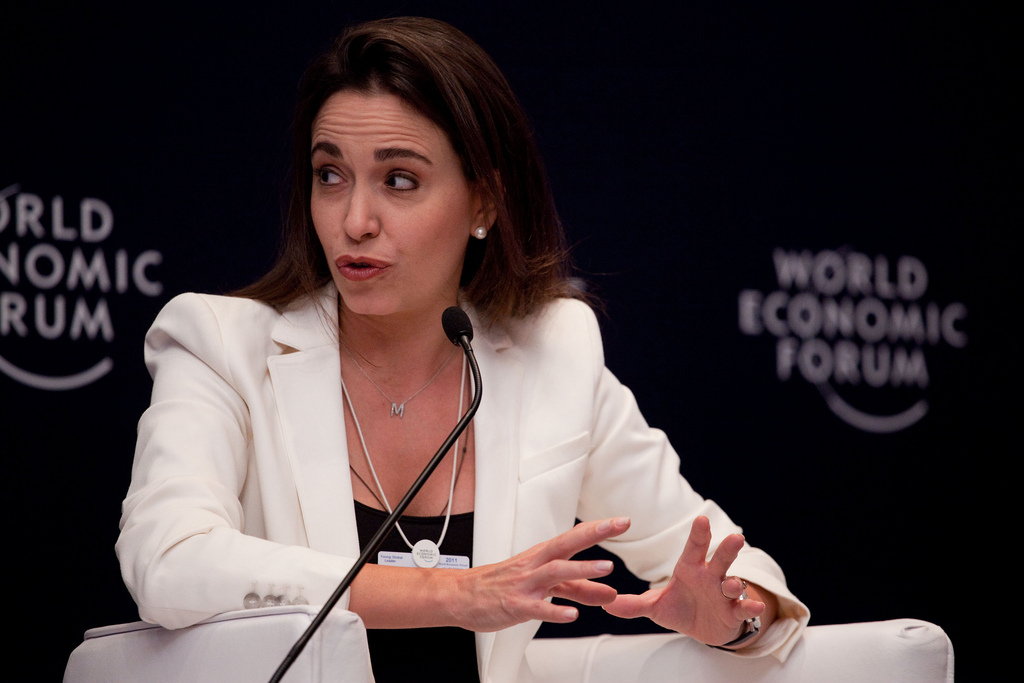 María Corina Machado, opposante vénézuélienne et lauréate du prix Nobel de la paix 2025, s’exprimant lors d’un panel du World Economic Forum, vêtue d’un tailleur blanc et parlant avec énergie devant un micro.